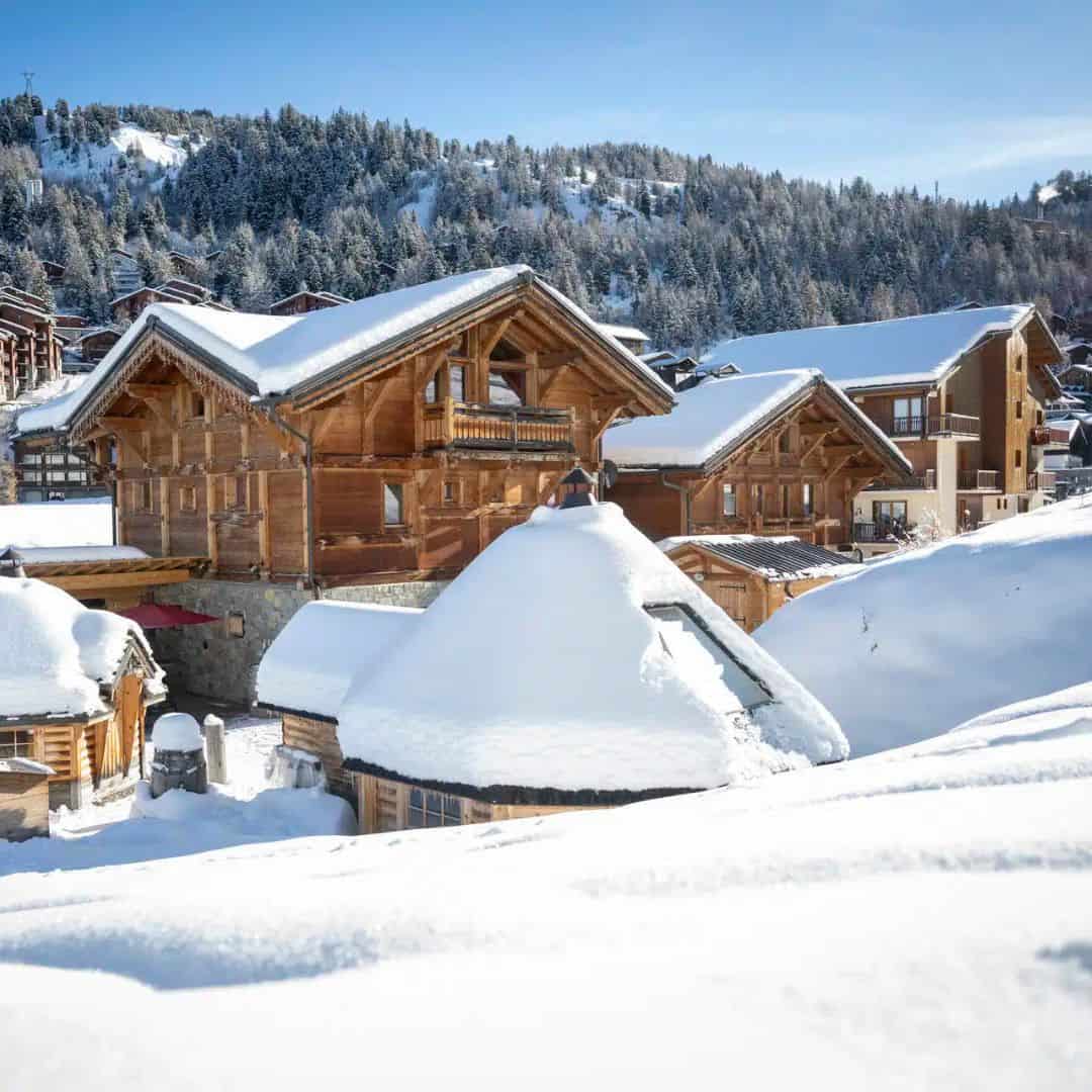 Charming wooden chalets with steep, snow-covered roofs nestled in a quiet mountain village. Pine trees blanket the hillside under a bright blue sky, giving a postcard-perfect alpine vibe.