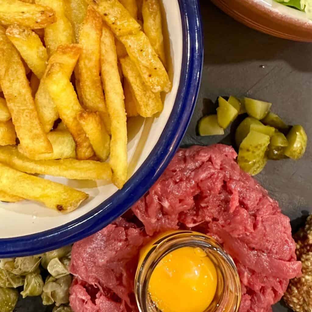 Close-up of steak tartare with a raw egg yolk in the center, surrounded by pickles and capers, with a bowl of seasoned French fries placed beside it.