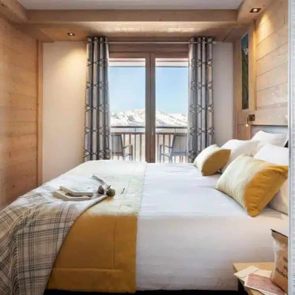 Bright bedroom with a large bed dressed in white and mustard yellow linens, facing a balcony that overlooks snowy mountain peaks. Flanked by patterned curtains and soft lighting, the space feels both inviting and luxurious.