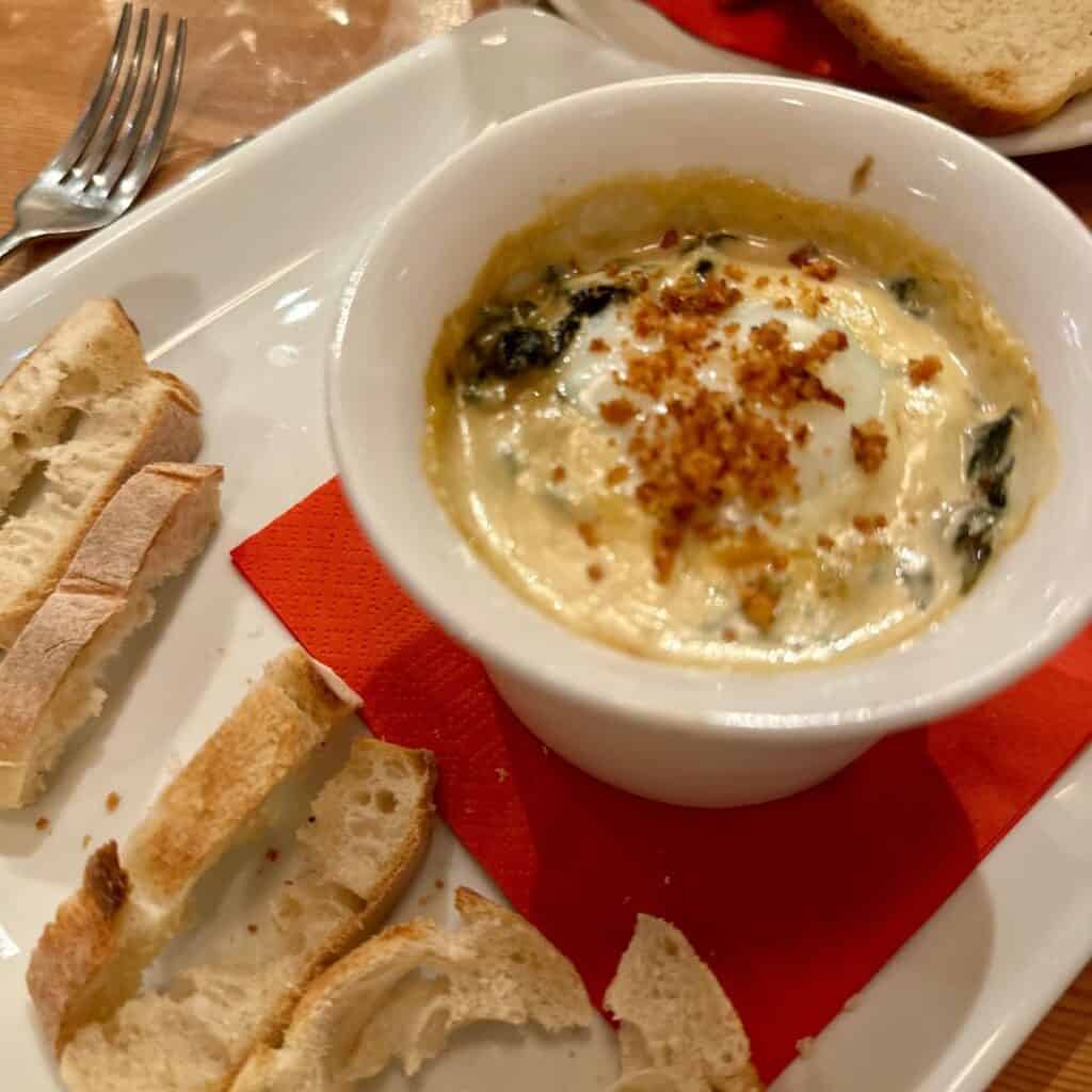 A bowl of creamy spinach and cheese dip topped with crispy breadcrumbs, served on a tray with slices of crusty baguette for dipping.