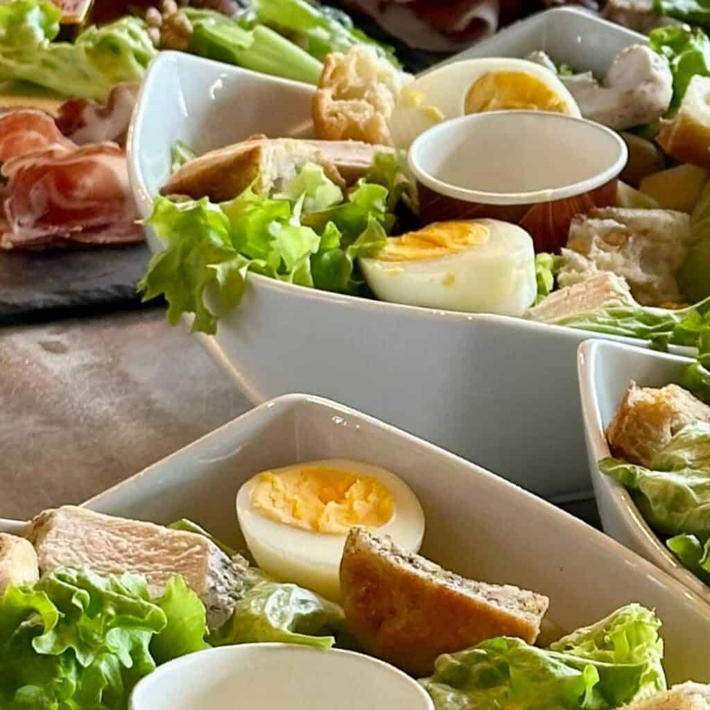 Fresh salads served in white bowls, each topped with crisp lettuce, boiled egg halves, croutons, and chicken, with a small cup of dressing nestled in the greens.