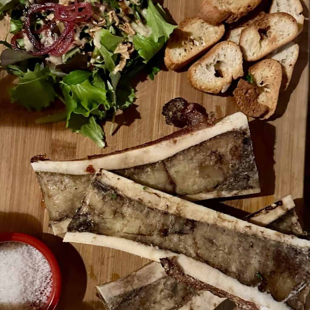 Overhead view of bone marrow bones on a wooden board, accompanied by salad greens, red onion, toasted bread, and a red ramekin of salt.
