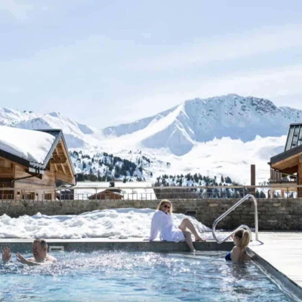 Three people enjoy a steamy outdoor pool surrounded by snow, with rustic wooden chalets and dramatic alpine peaks in the background. Clear skies and sunshine contrast with the icy landscape.