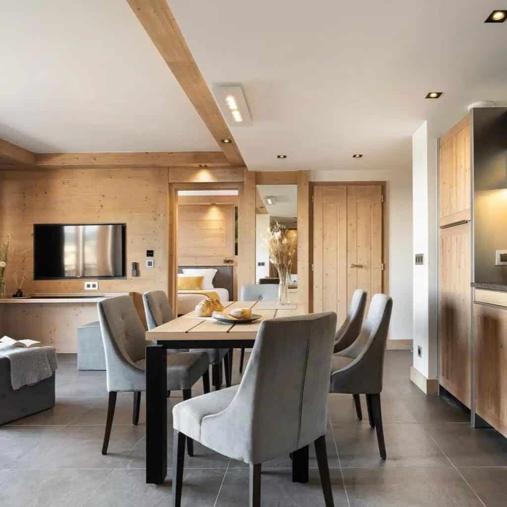 Elegant dining area with plush gray chairs and a wooden table set for a meal, connected to a stylish living room and kitchen. Light wood accents and soft lighting create a welcoming, modern alpine vibe.