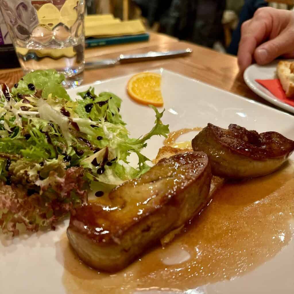 Seared foie gras drizzled in a rich sauce served alongside a fresh green salad with balsamic glaze and a slice of orange for garnish.