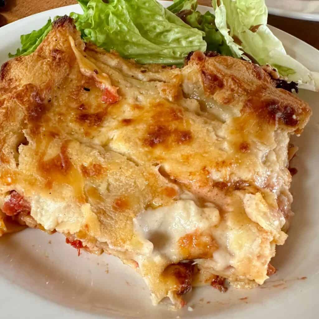 Close-up of a golden-baked slice of cheesy lasagna with visible layers of pasta, sauce, and béchamel, served with a side of fresh green lettuce on a white plate.