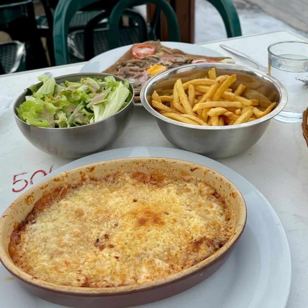 A white tabletop holds a hearty baked dish, likely lasagna or gratin, alongside metal bowls filled with French fries and a green salad. In the background, a buckwheat galette topped with sliced tomato, an egg, and meat is partially visible.