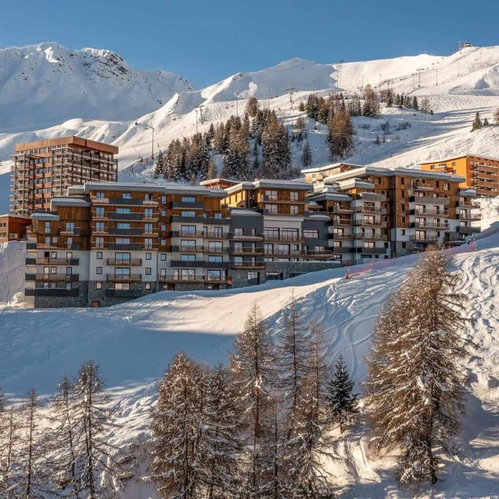 Wide view of a modern alpine ski resort nestled in the snowy mountains, with multi-story chalet-style buildings and pine trees scattered along the slope under bright winter sun.