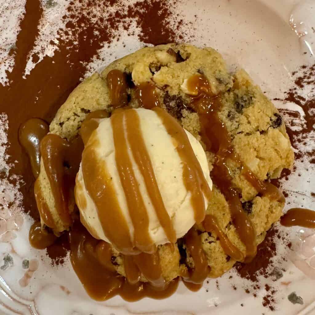A warm chocolate chip cookie topped with a scoop of vanilla ice cream and drizzled generously with caramel sauce, served on a dessert plate dusted with cocoa powder.