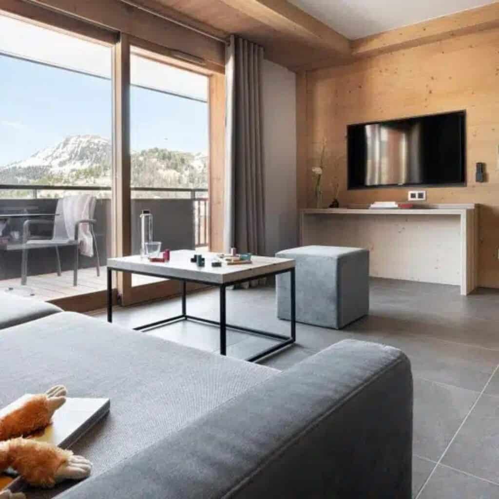 Cozy living room with a gray sectional sofa, coffee table, and wall-mounted TV, opening to a balcony with clear mountain views. The room features warm wood paneling, soft lighting, and minimalist decor.