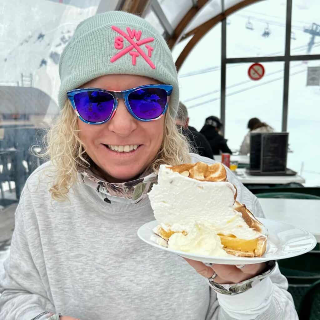 A woman in blue mirrored sunglasses and a light green beanie with pink "SWT" letters smiles while holding a large waffle topped with whipped cream and custard. She's seated indoors at a ski lodge with snow and chairlifts visible through the windows behind her.