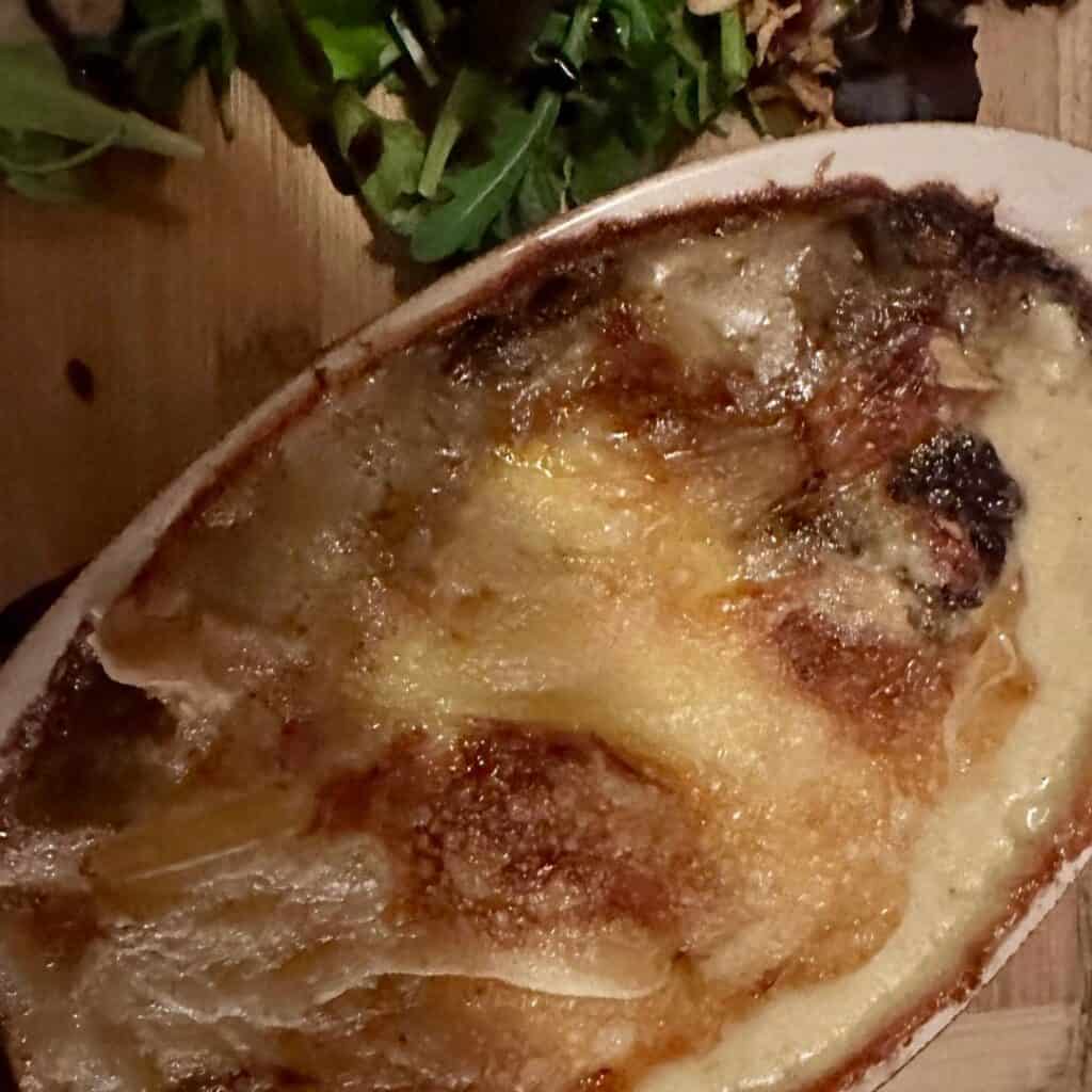 A bubbling gratin dish with a golden, crispy cheese topping, served alongside a small portion of mixed greens on a wooden board.