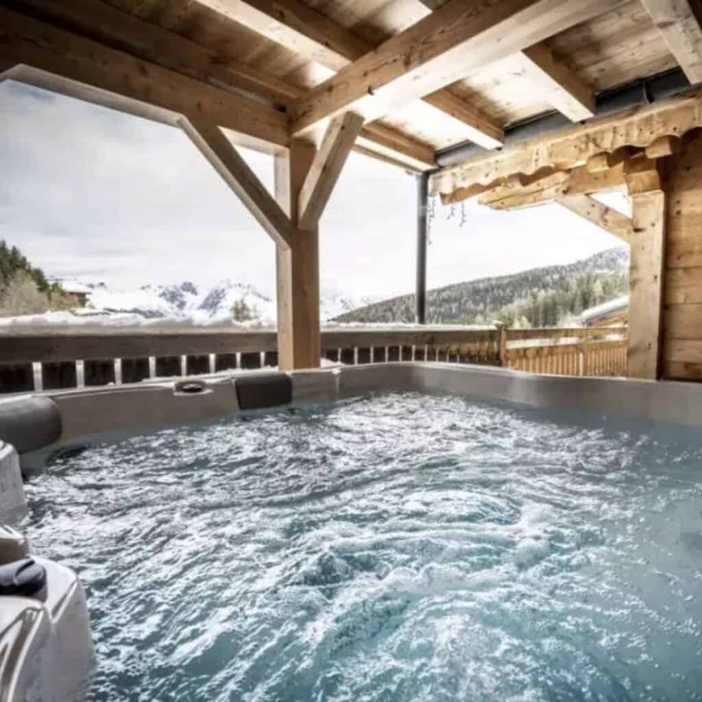 A bubbling hot tub on a wooden deck under a covered roof, overlooking snow-dusted pine forests and distant mountains. The space blends privacy with natural beauty, perfect for après-ski relaxation.
