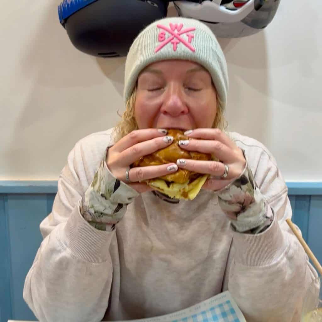 A woman in a light sweatshirt and beanie eagerly takes a bite of a large burger, eyes closed in enjoyment, seated in a cozy restaurant setting.