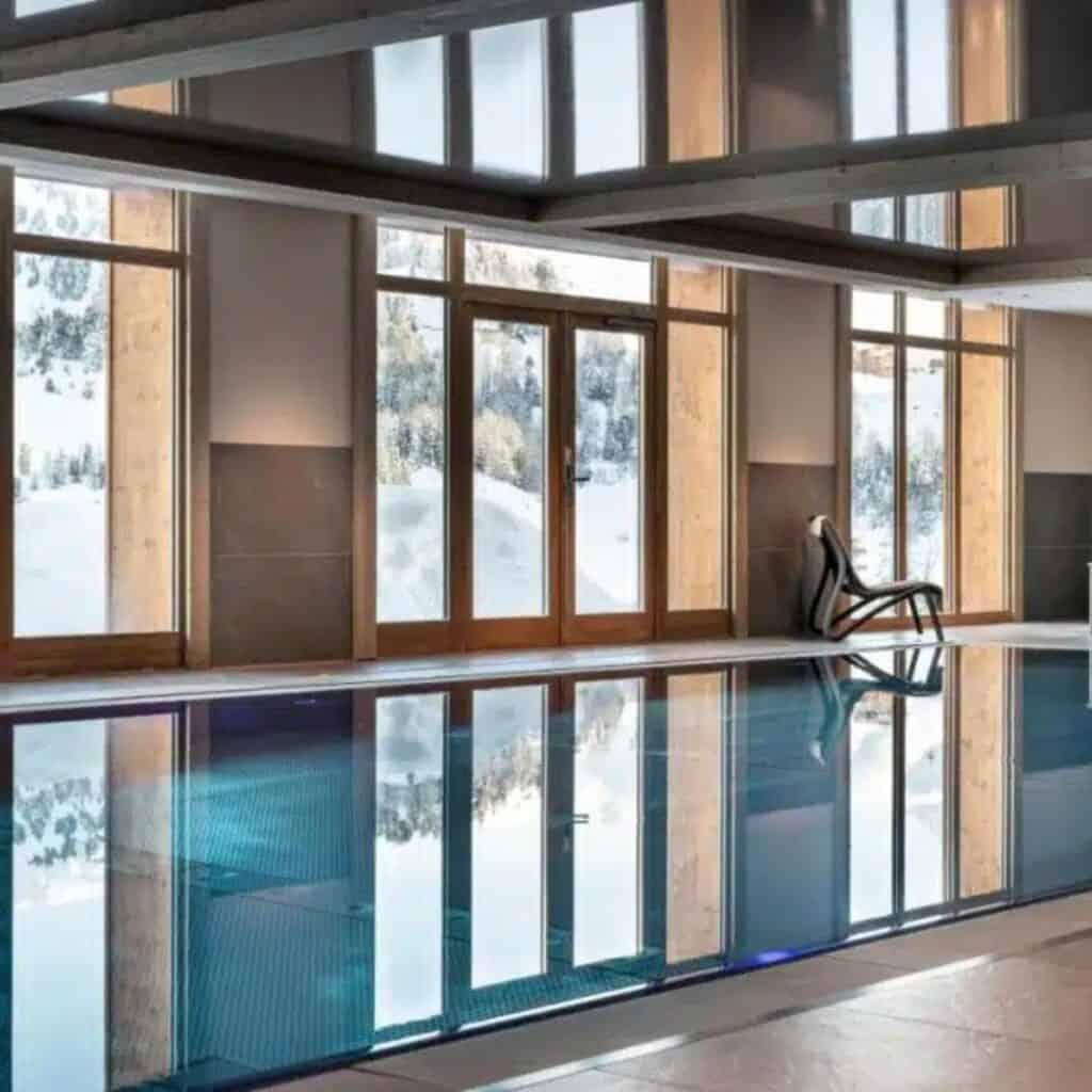 Modern indoor swimming pool with sleek lounge chairs and floor-to-ceiling windows showcasing a snowy mountain landscape. The still water reflects the wood-framed glass panels and alpine scenery outside.