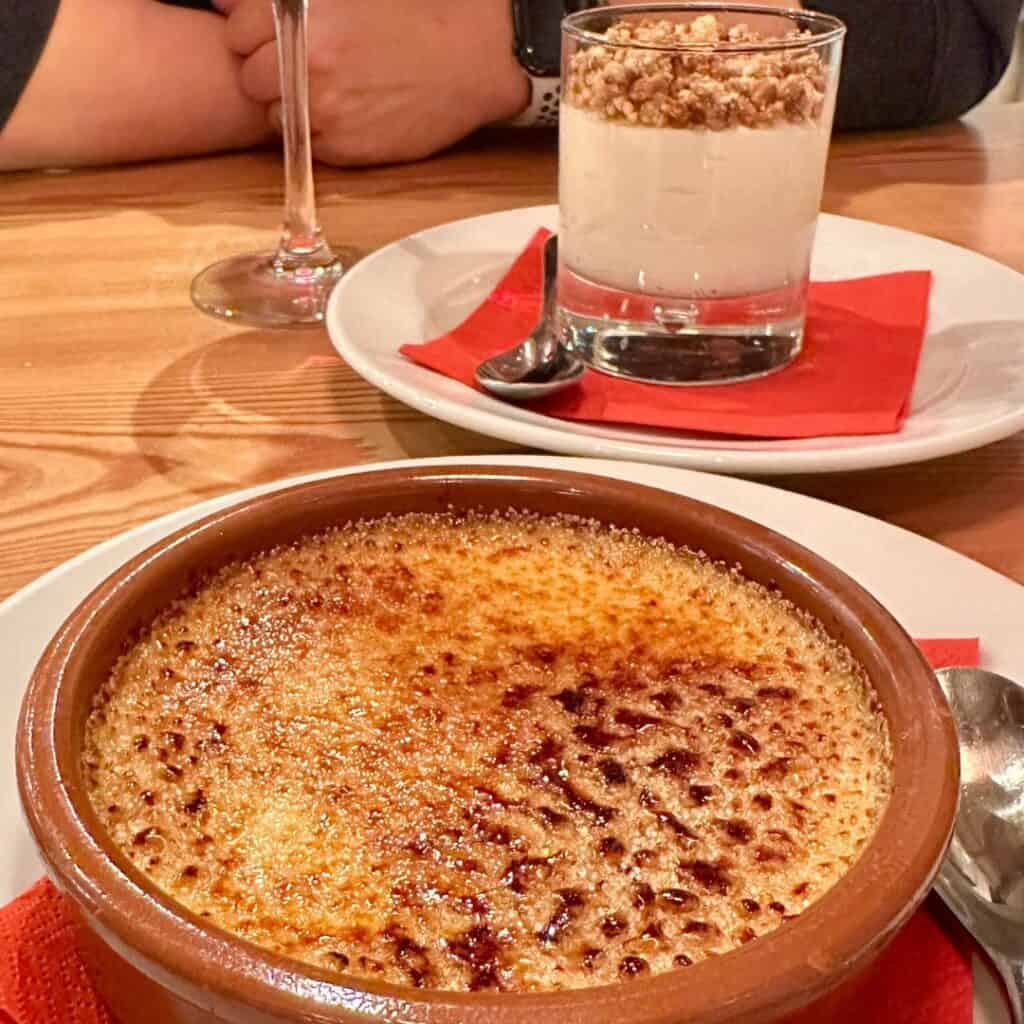 Classic crème brûlée with a golden, caramelized sugar crust served in a rustic ceramic dish, with a creamy parfait topped with crunchy granola in the background.