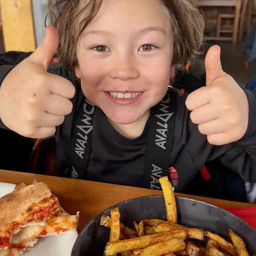 Smiling child gives two enthusiastic thumbs up while sitting in front of a bowl of golden fries and a slice of cheesy pizza, clearly loving the meal.