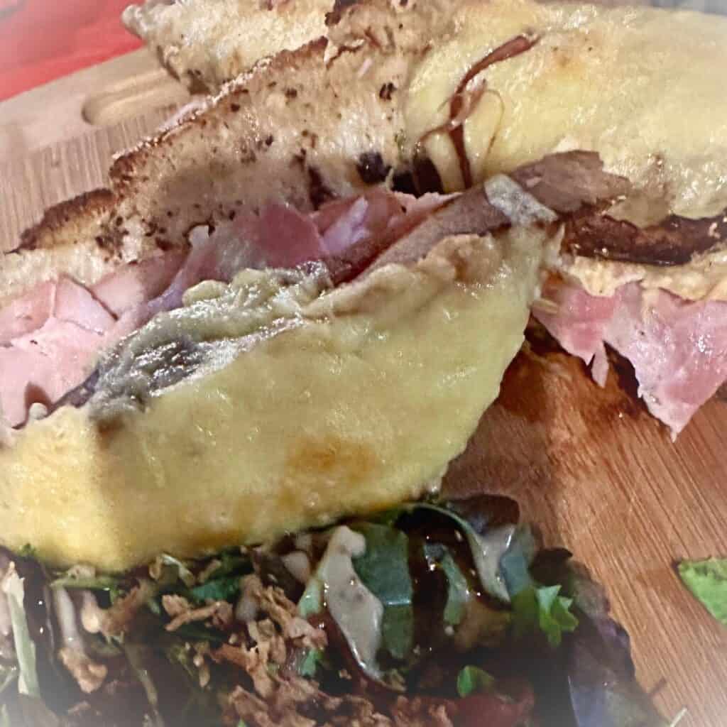 A warm ham and cheese toastie made with crusty bread, melted cheese oozing over the edges, served with a side salad on a wooden board.