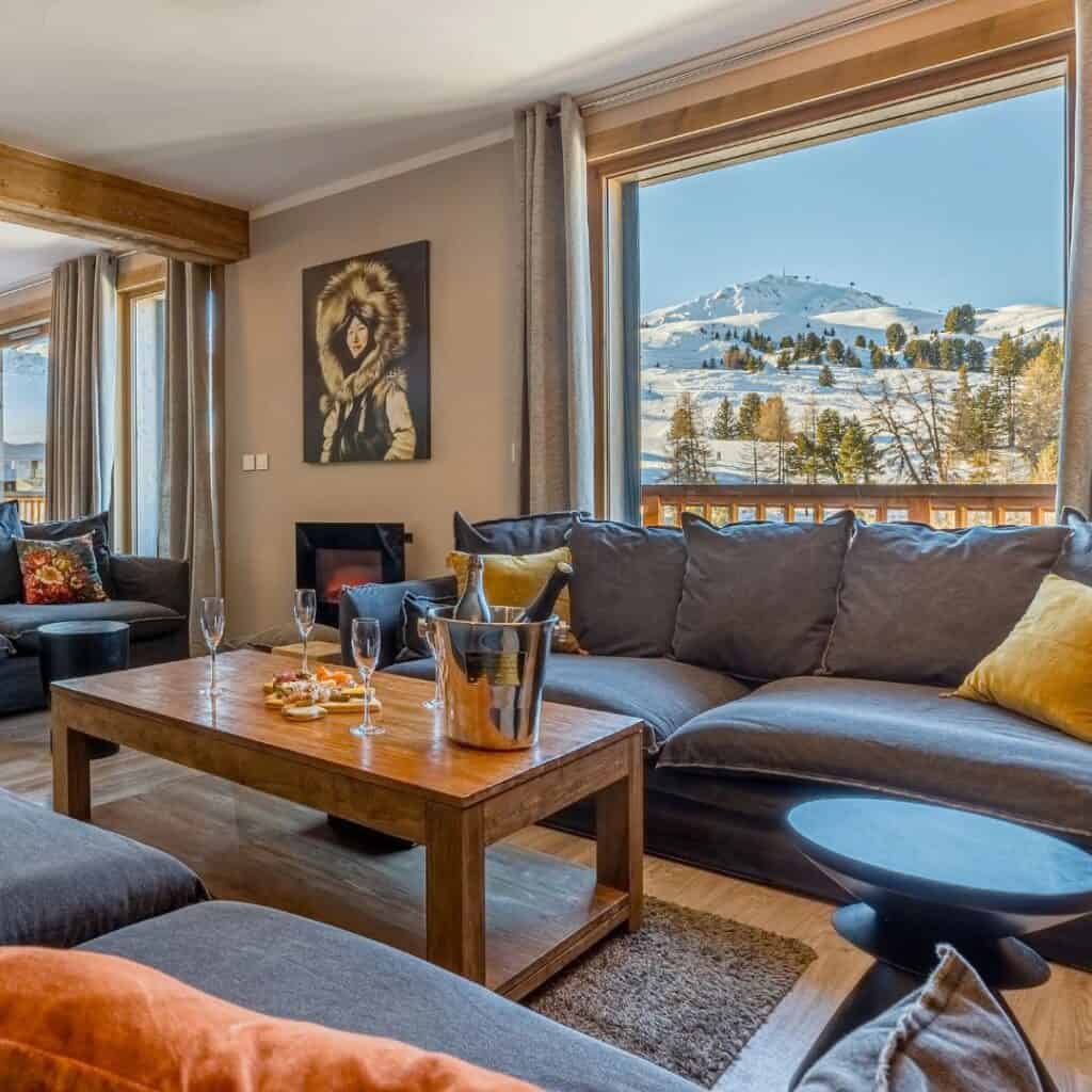 Warm and inviting living room with plush sofas, a wooden coffee table set with champagne and snacks, and panoramic views of snowy slopes through large windows. Decor includes earthy tones and a striking portrait of a woman in winter gear.