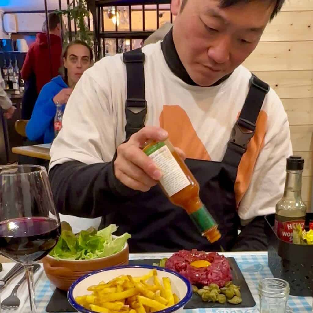 A man in ski overalls adds hot sauce to a plate of steak tartare topped with a raw egg yolk, served with capers, pickles, a bowl of fries, and salad at a gingham-covered table.