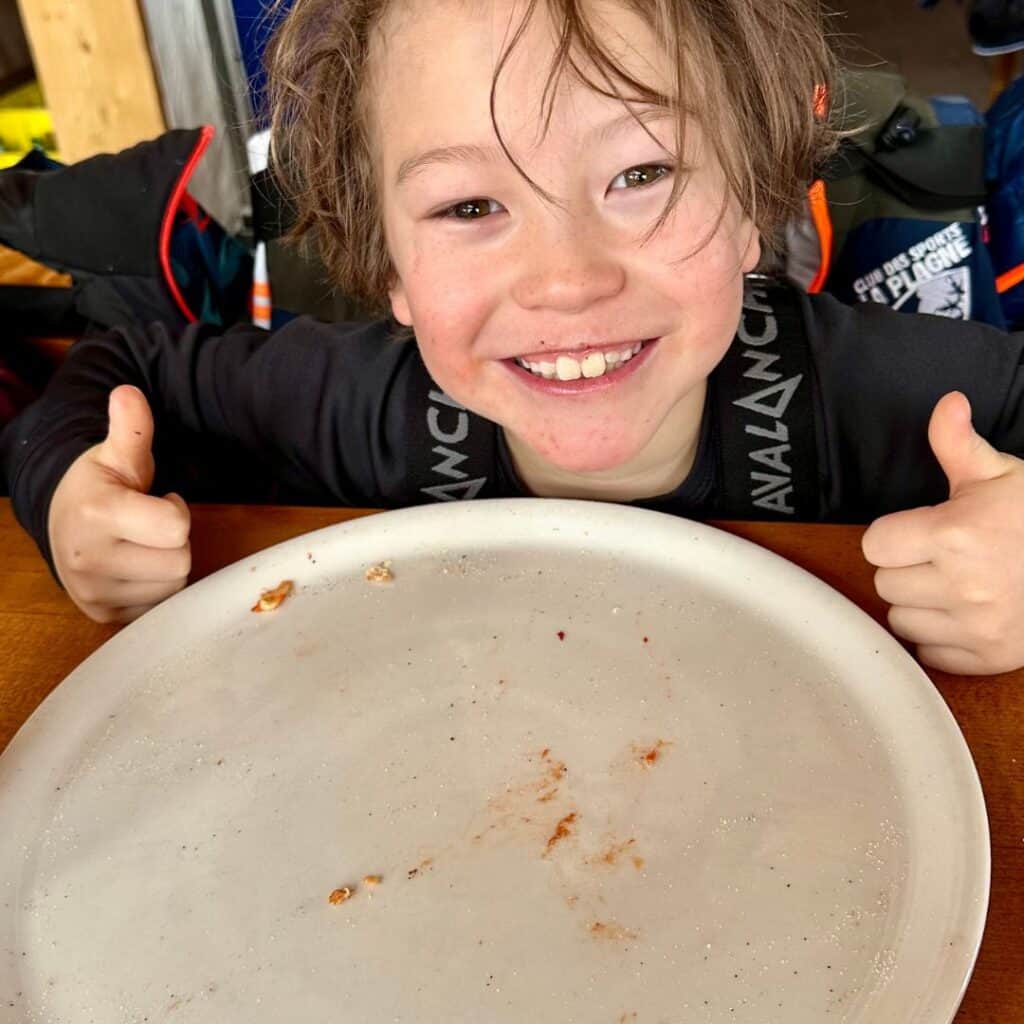 Grinning child gives two thumbs up in front of an empty white plate with just a few sauce smudges left, showing a meal thoroughly enjoyed.
