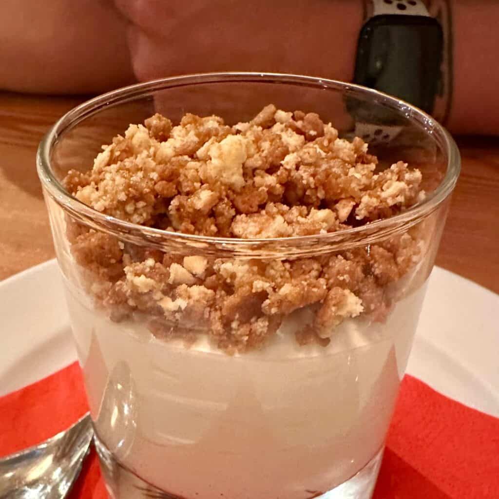 Close-up of a glass parfait layered with creamy yogurt and finished with a thick crumble topping, placed on a red napkin.