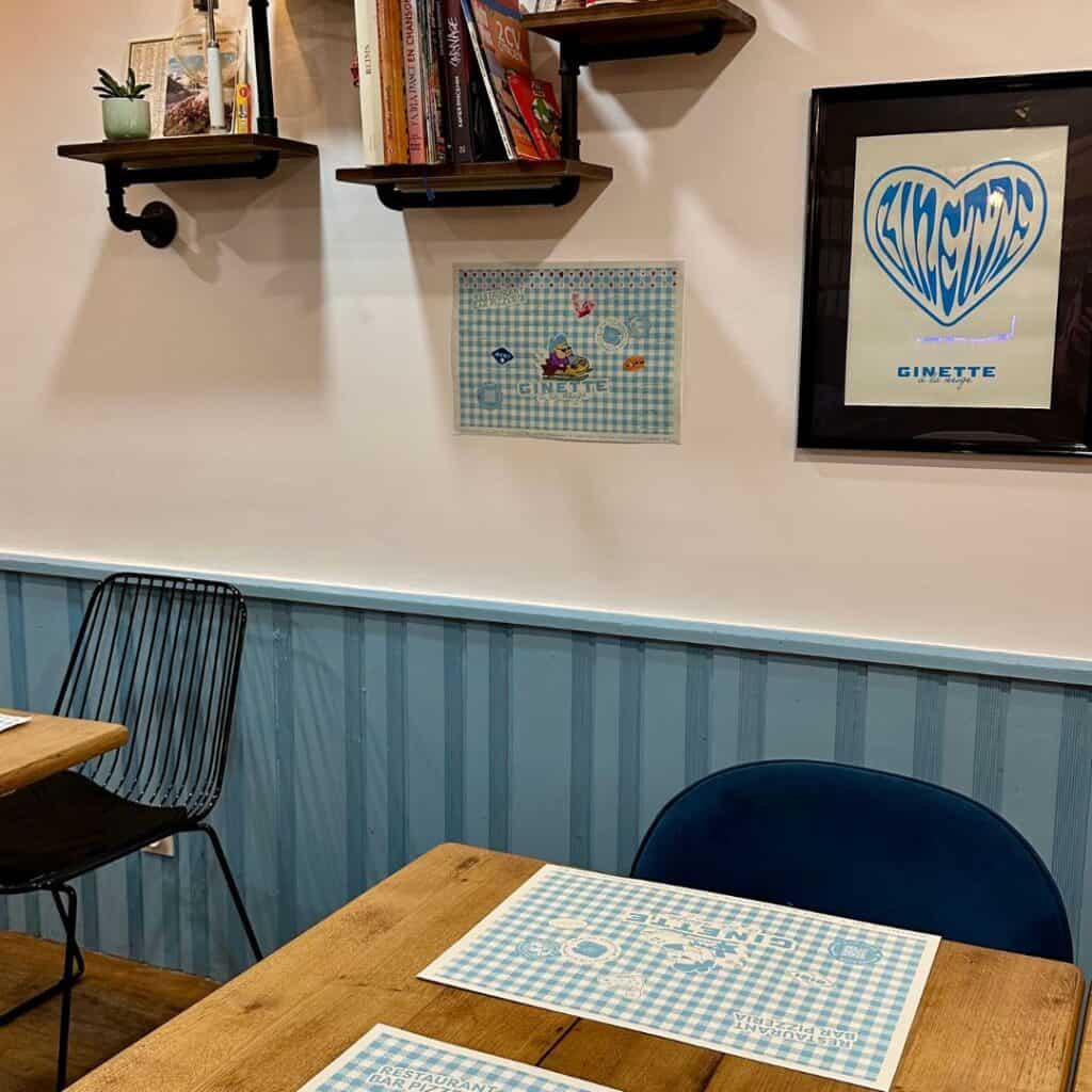 Cozy restaurant interior featuring wooden tables set with blue gingham placemats labeled “Ginette.” The light blue wainscoting and white walls are decorated with framed artwork and floating shelves holding books and small plants. Simple wire and cushioned chairs complete the casual, charming vibe.