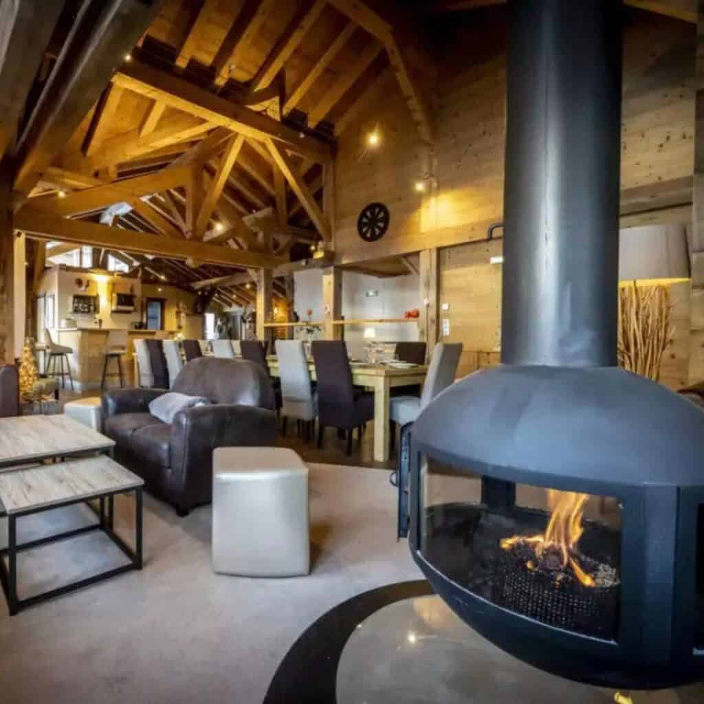 Rustic-modern chalet living space featuring exposed wooden beams, cozy lounge seating, and a central black fireplace. The warm glow of the fire and ambient lighting enhances the inviting atmosphere.