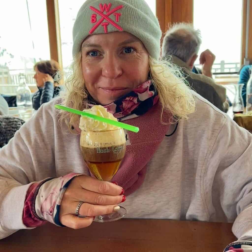 Woman in a grey beanie and pink scarf smiles while holding a glass of Irish coffee topped with whipped cream and a green straw, seated inside a cozy restaurant, Le Carroley in La Plagne.