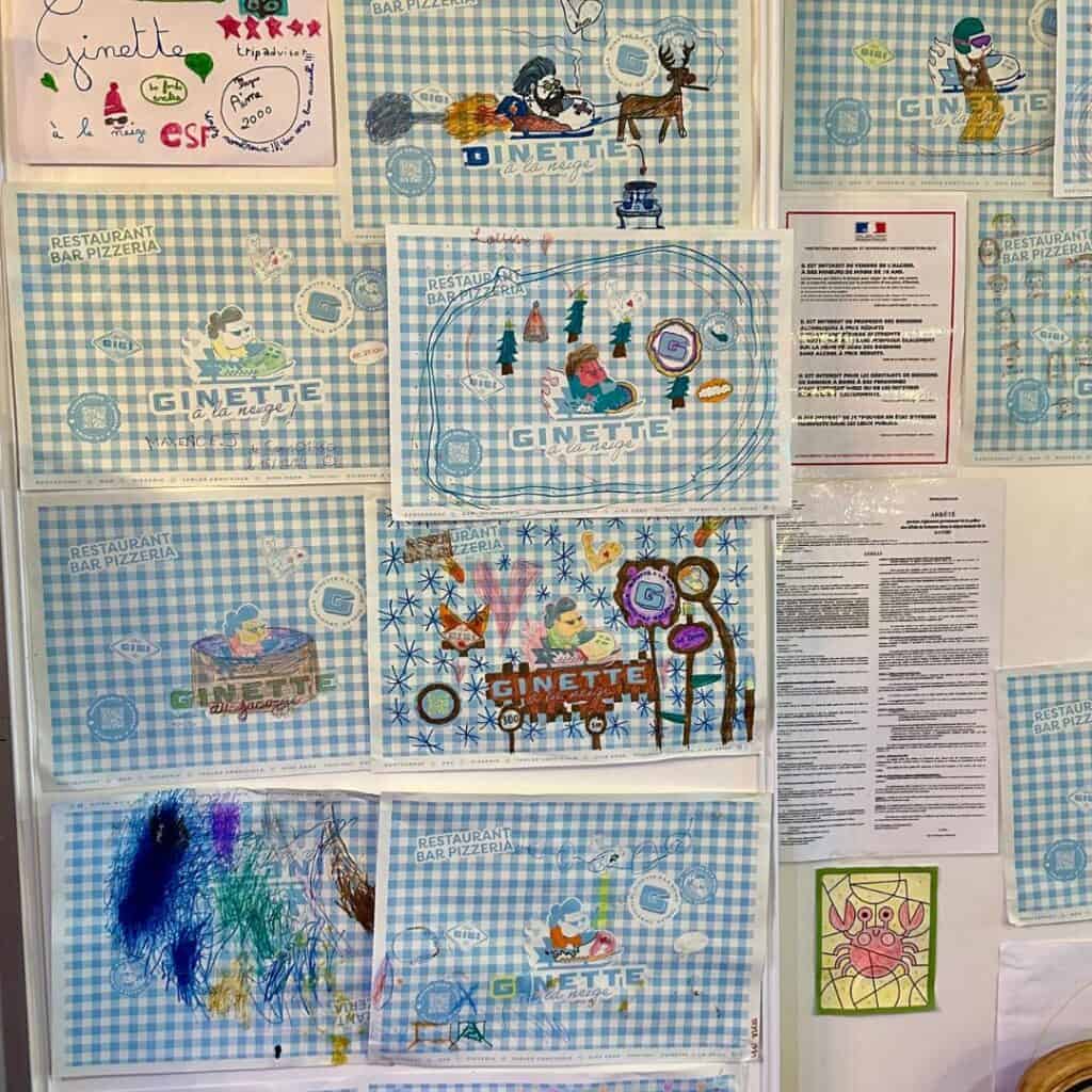 A wall display filled with blue gingham Ginette restaurant placemats creatively colored and decorated by children. Each placemat features unique drawings like animals, mountains, and skiers, adding a playful and personal touch to the restaurant’s cozy atmosphere. French notices and a few additional drawings are posted in between.