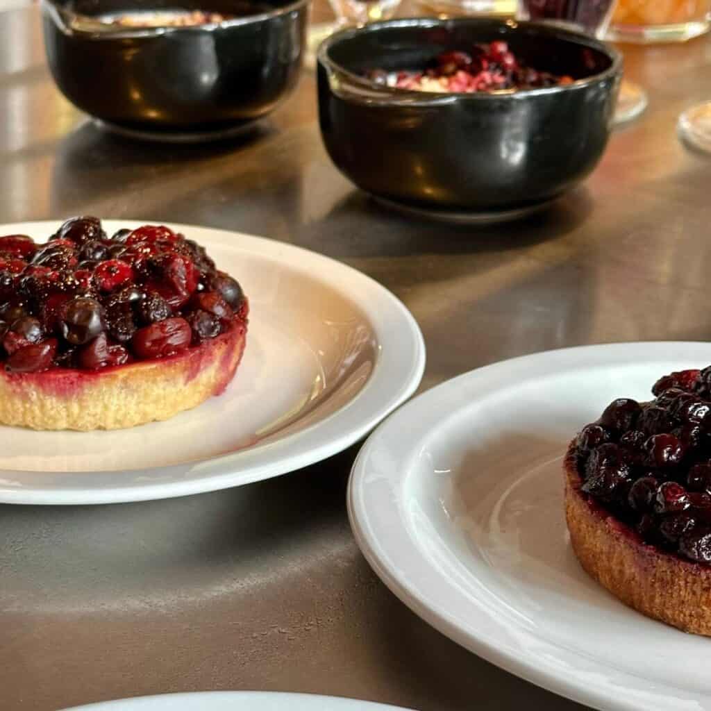 Small berry tarts on white plates topped with glossy black and red berries, surrounded by black bowls filled with other desserts on a metal tabletop.