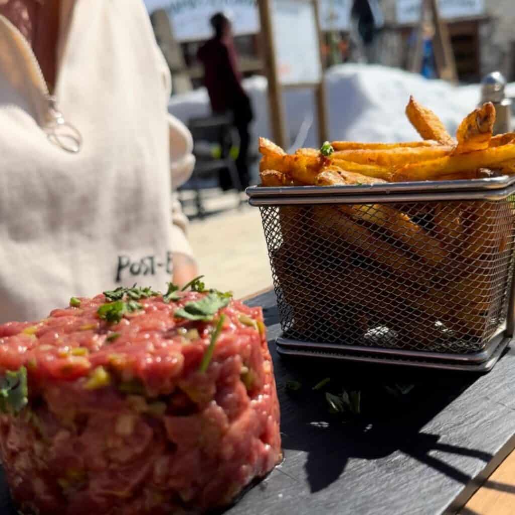 A serving of steak tartare garnished with fresh herbs is plated beside a metal basket of golden, crispy French fries. The meal is presented on a slate board outdoors, with sunshine casting shadows and a casual dining atmosphere in the background.