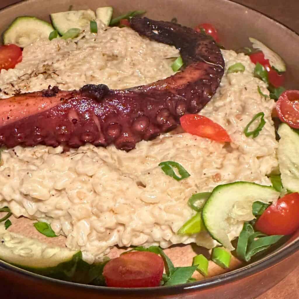 Creamy risotto served in a bowl, topped with a grilled octopus tentacle and garnished with fresh cucumber slices, halved cherry tomatoes, and chopped scallions. The dish offers a rich and colorful presentation with a Mediterranean flair.