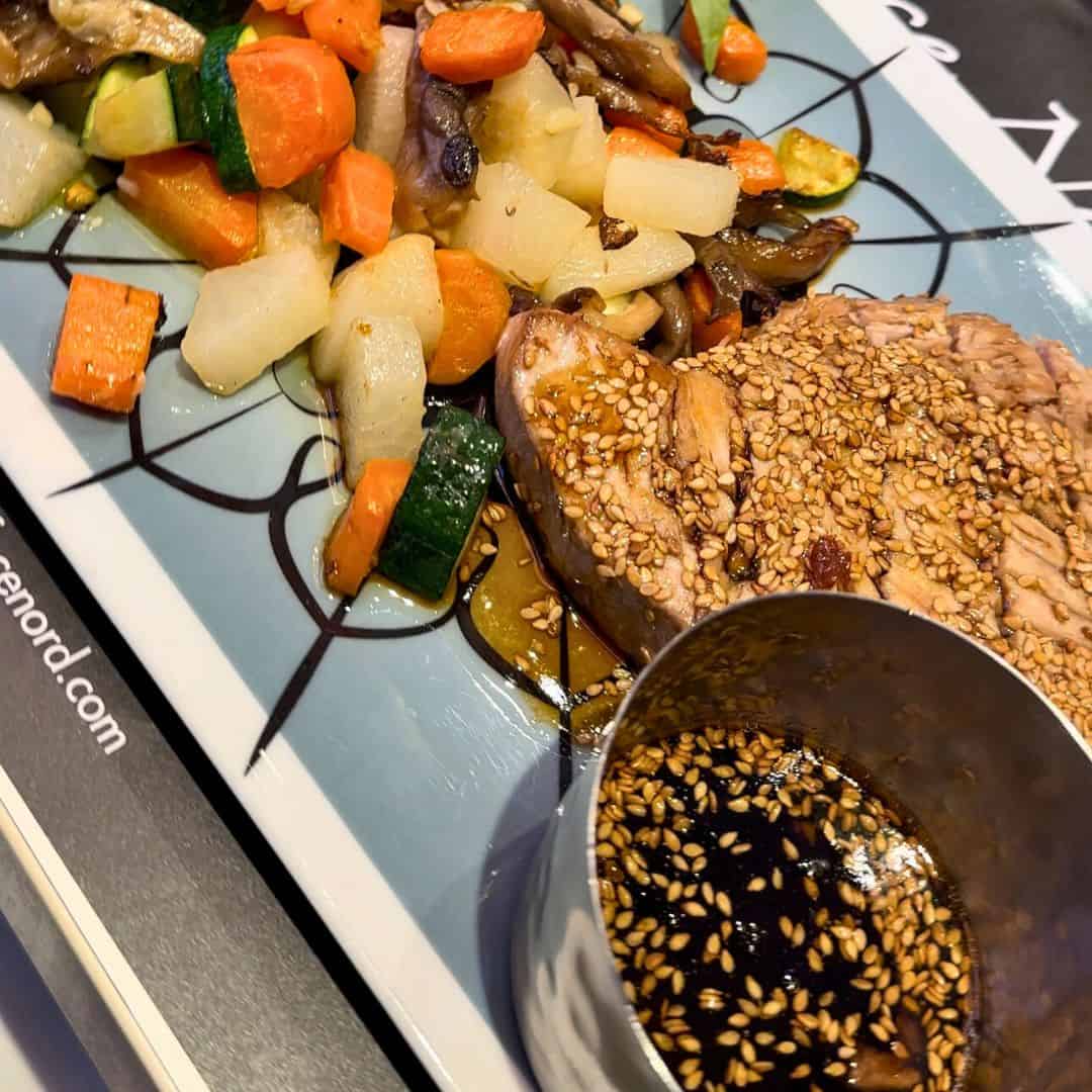 Plate of sesame-crusted tuna steak served with a medley of sautéed vegetables including carrots, zucchini, and mushrooms, alongside a small metal cup of soy-based dipping sauce topped with sesame seeds. The colorful presentation sits on a modern, patterned dish.