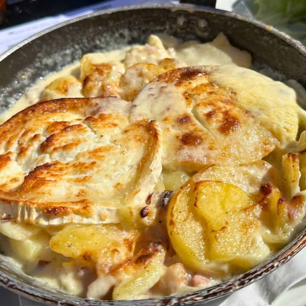 Close-up of a hearty tartiflette served in a skillet, featuring golden-brown potato slices baked in a creamy cheese sauce with melted Reblochon on top. The rich dish includes visible bits of bacon and onions, capturing a warm, comforting alpine meal.