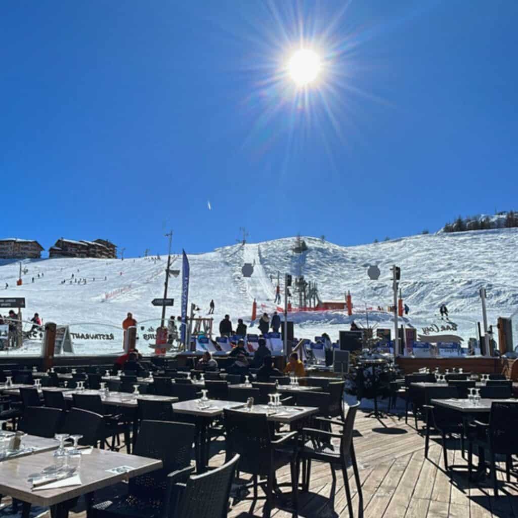 A sunny ski resort scene with an outdoor dining terrace in the foreground, set with black wicker chairs and tables under clear blue skies. In the background, skiers glide down a snow-covered slope with ski lifts and chalets visible atop the hill.