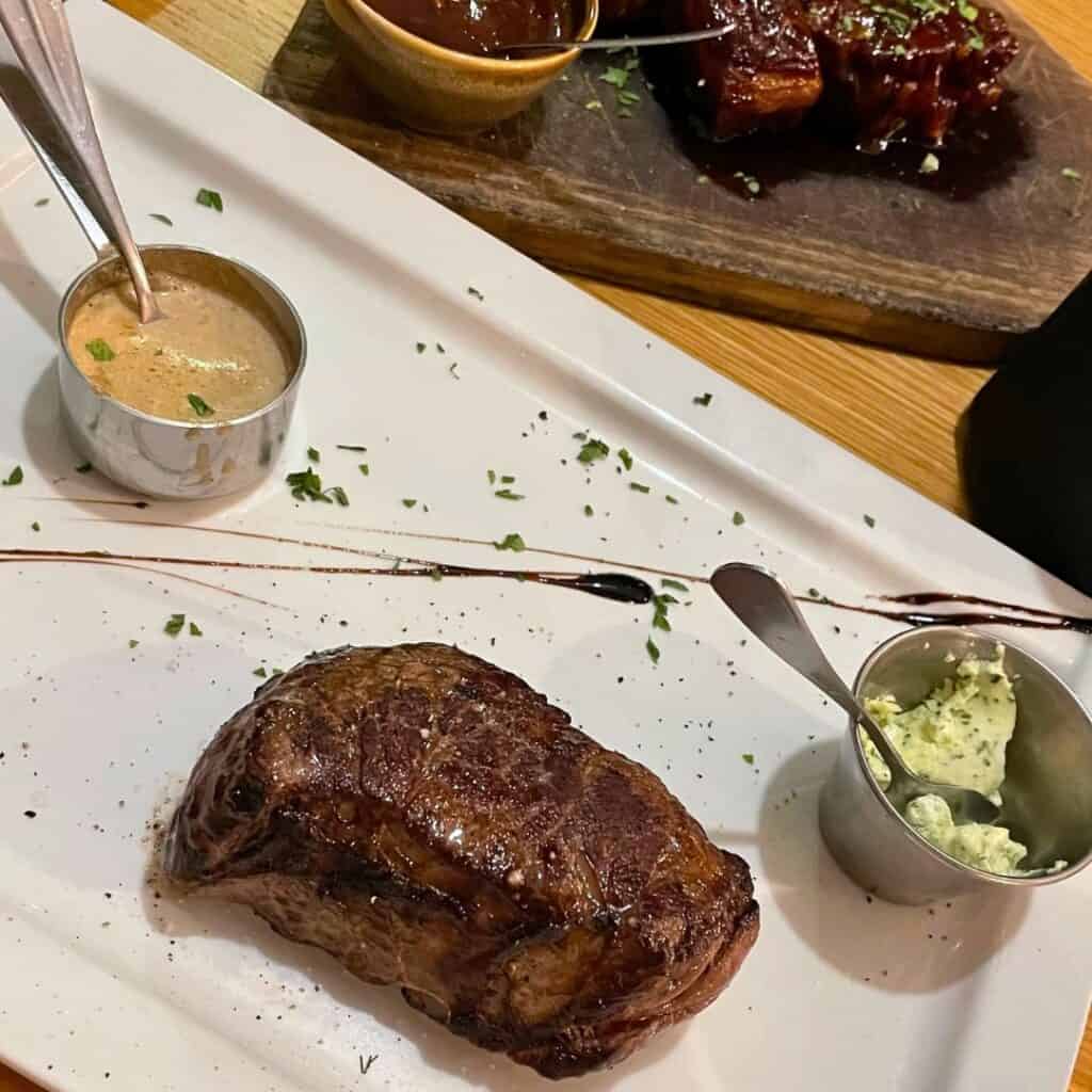 A perfectly seared steak served on a white rectangular plate, accompanied by two small metal cups of creamy peppercorn sauce and herbed butter, with a balsamic glaze drizzle and chopped parsley for garnish. In the background, a wooden board holds glazed ribs and an extra ramekin of sauce.