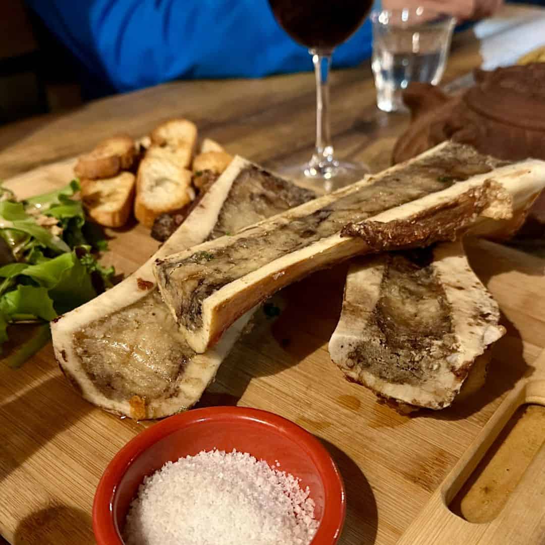 Close-up of roasted bone marrow presented on a wooden board with coarse salt, salad, and toasted bread in the background.