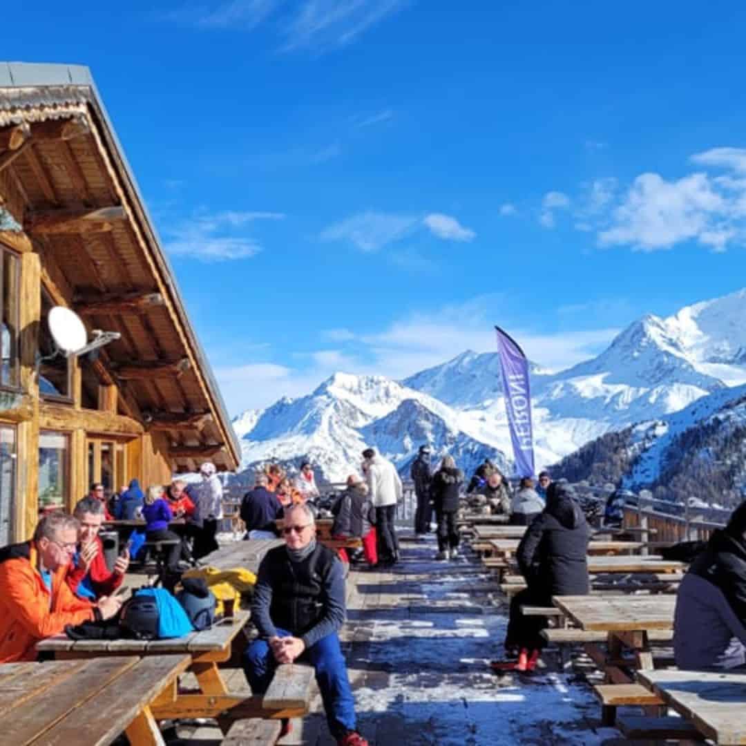 People relaxing at outdoor wooden picnic tables beside a rustic mountain lodge, with snowy peaks and a bright blue sky in the background. A purple flag reading “La Rosière” flutters among the crowd, suggesting a ski resort setting in the French Alps. Snow patches cover the wooden deck, and many visitors are dressed in winter gear, enjoying the sunshine.