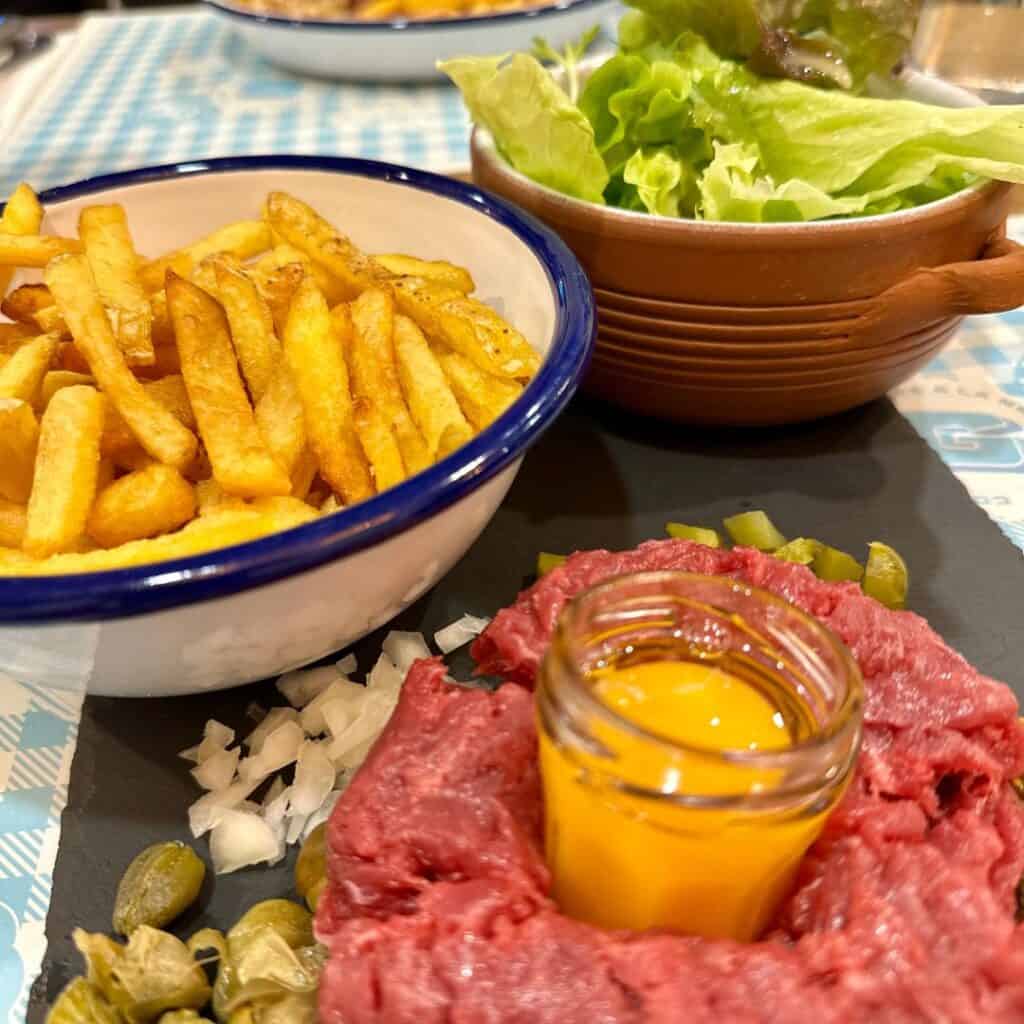 Classic steak tartare served with a raw egg yolk in a glass shot glass at the center, surrounded by chopped onions, capers, and pickles. The dish is paired with a bowl of golden French fries and a side salad with fresh lettuce in a ceramic bowl.