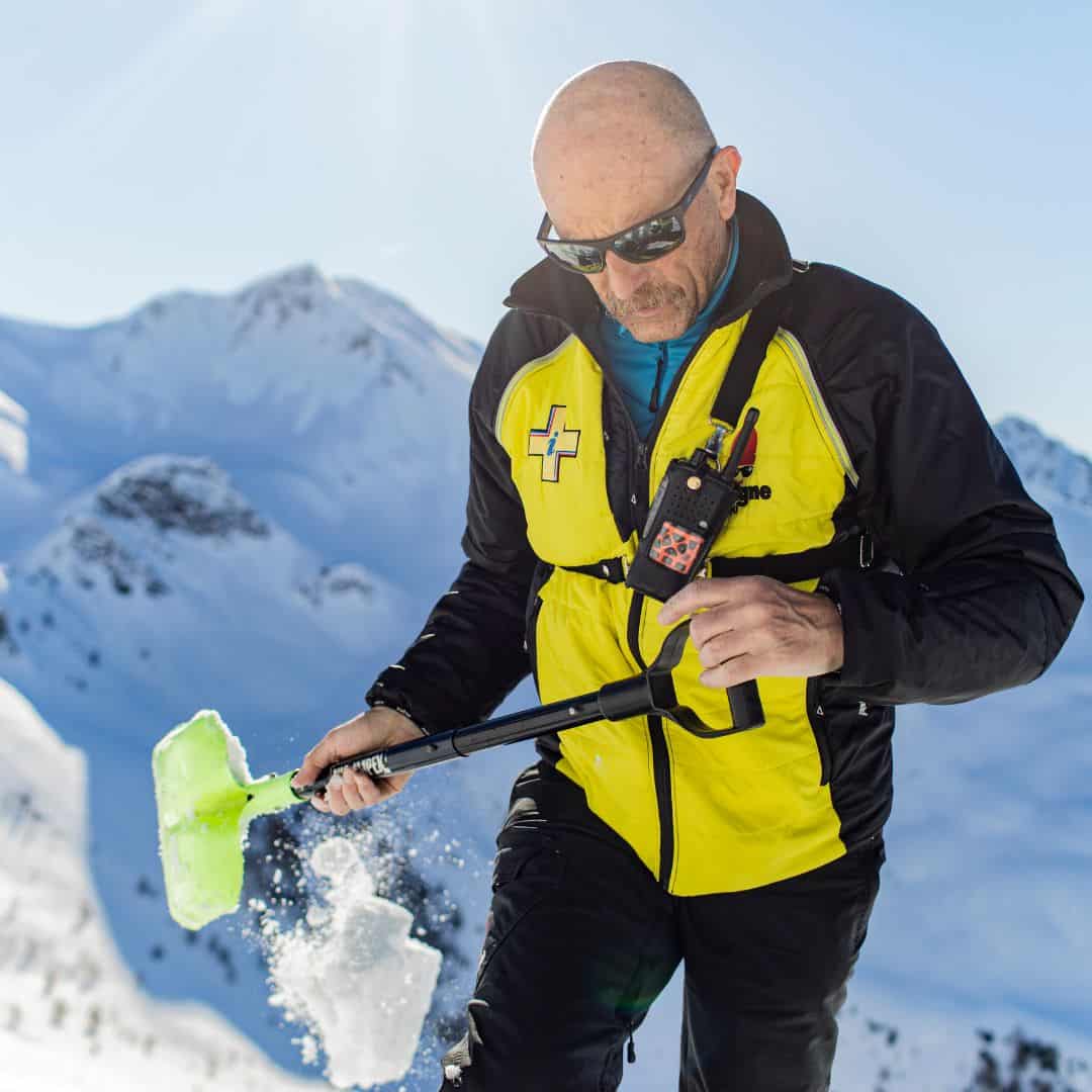 A ski patroller wearing a yellow and black jacket with a rescue insignia uses a shovel to examine snow conditions on a sunny mountain slope. He wears sunglasses and carries a radio, with snow-capped peaks in the background.