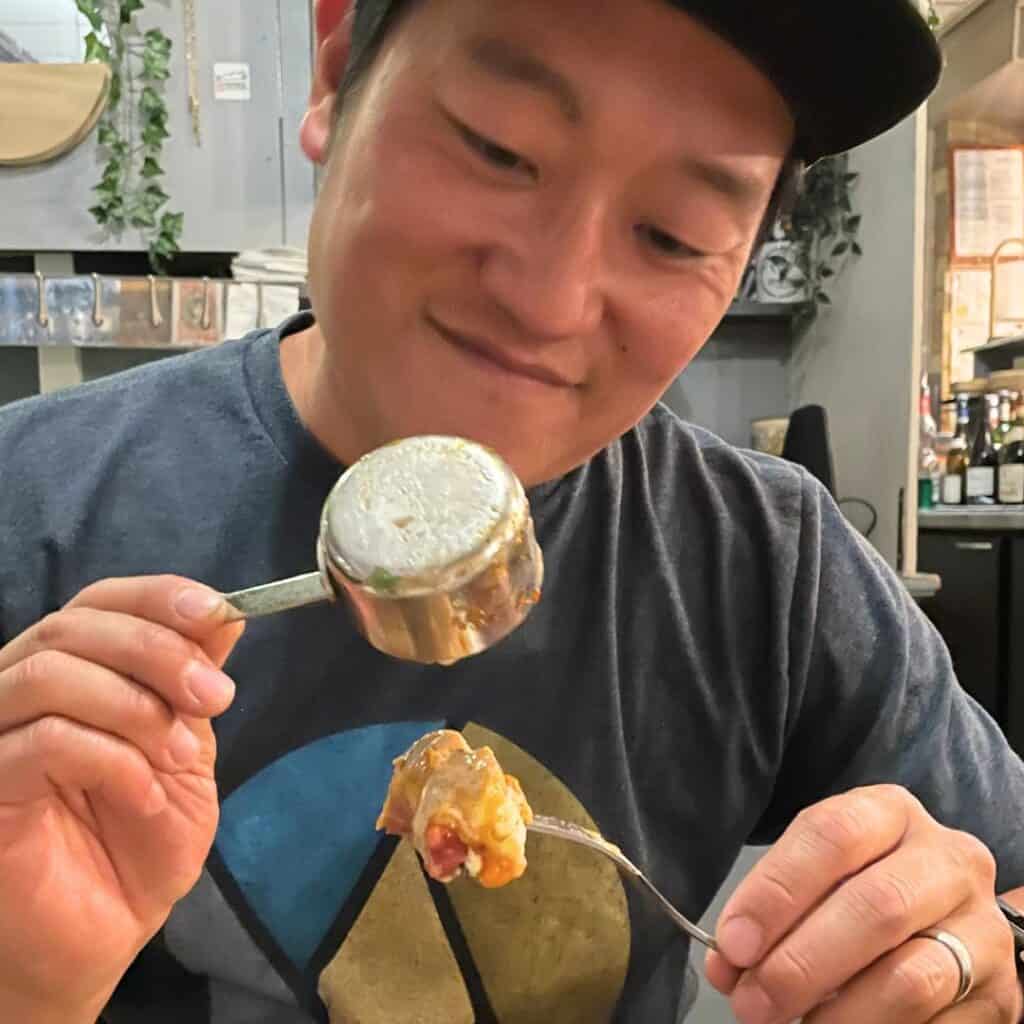 A smiling man in a casual t-shirt and cap holds up a small metal ramekin and a forkful of cheesy, savory food, ready to take a bite. The cozy restaurant setting in the background adds to the warm, relaxed vibe of the moment.