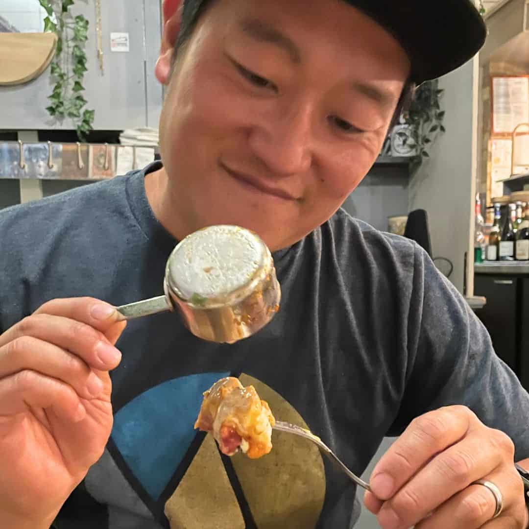 A smiling man in a casual t-shirt and cap holds up a small metal ramekin and a forkful of cheesy, savory food, ready to take a bite. The cozy restaurant setting in the background adds to the warm, relaxed vibe of the moment.