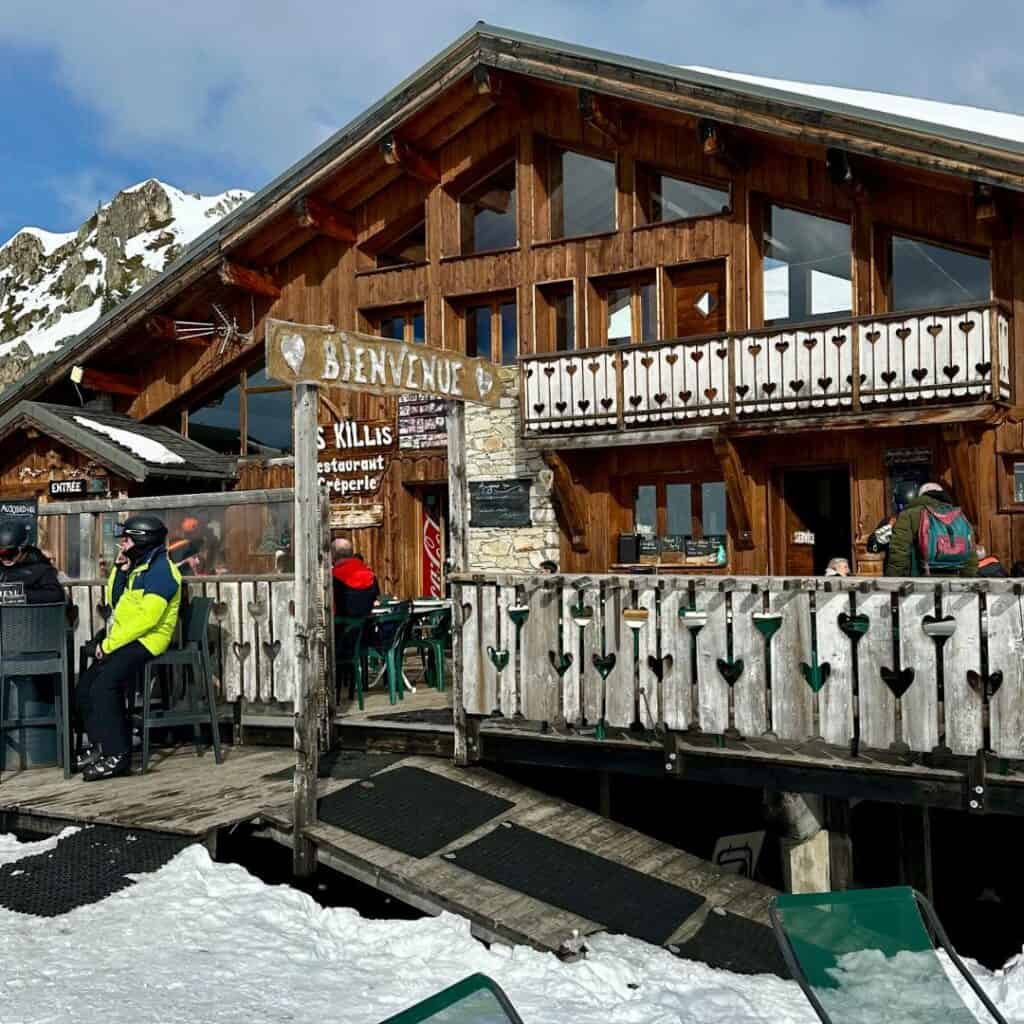 A rustic wooden chalet-style restaurant named “Les Killis” sits at the base of a snowy mountain, with skiers in helmets relaxing on the patio. A wooden sign reading “Bienvenue” welcomes visitors, and the building features heart-shaped cutouts on the railing and large windows reflecting the alpine scenery.