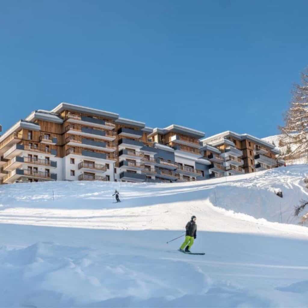 Skiers glide down a gentle snowy slope in front of a large, chalet-style apartment complex with wooden balconies, set against a clear blue sky at a ski resort.