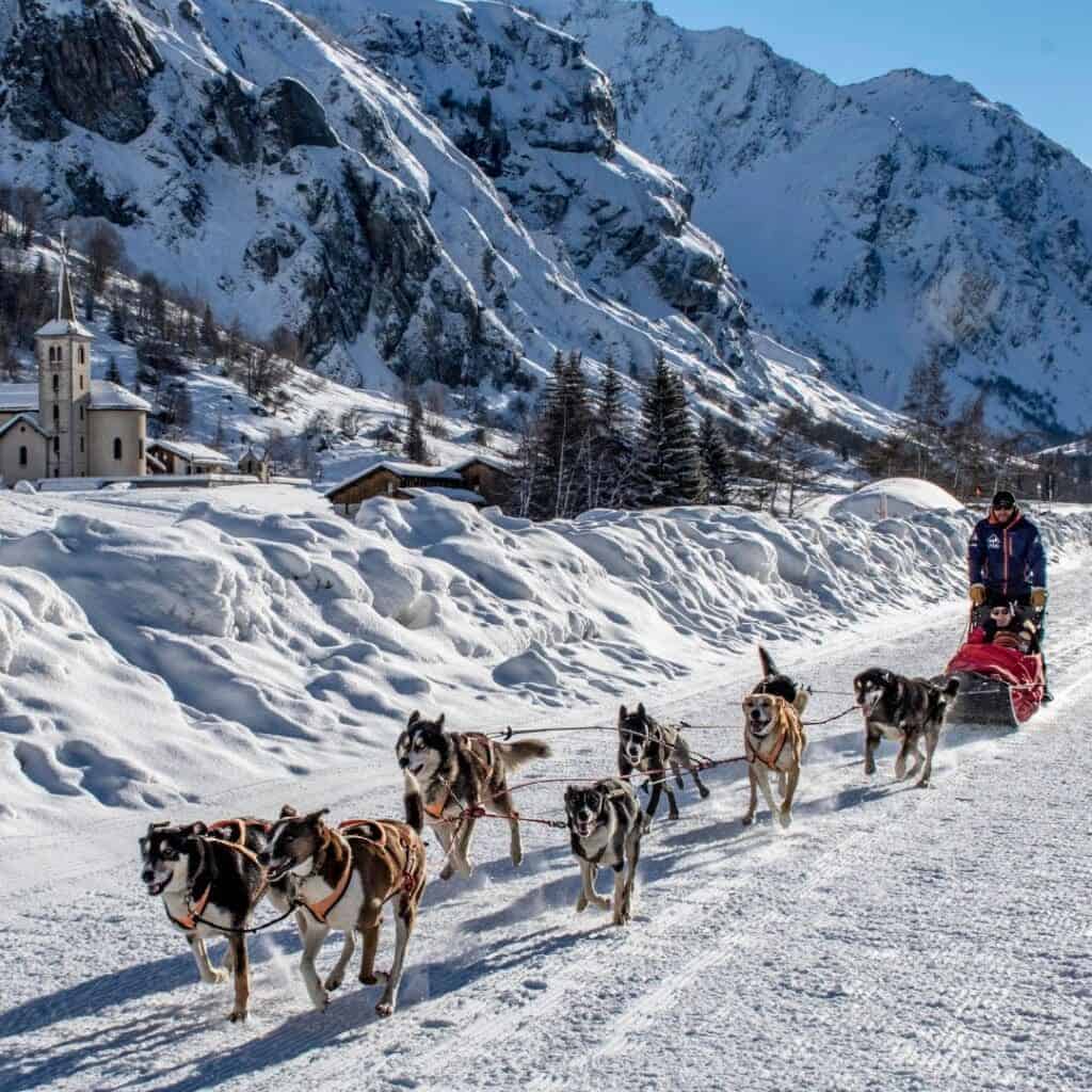 A team of huskies pulls a sled with two people through a snowy alpine valley, surrounded by towering snow-covered mountains and a small village with a church in the distance. The sky is clear and blue, adding to the stunning winter scenery.