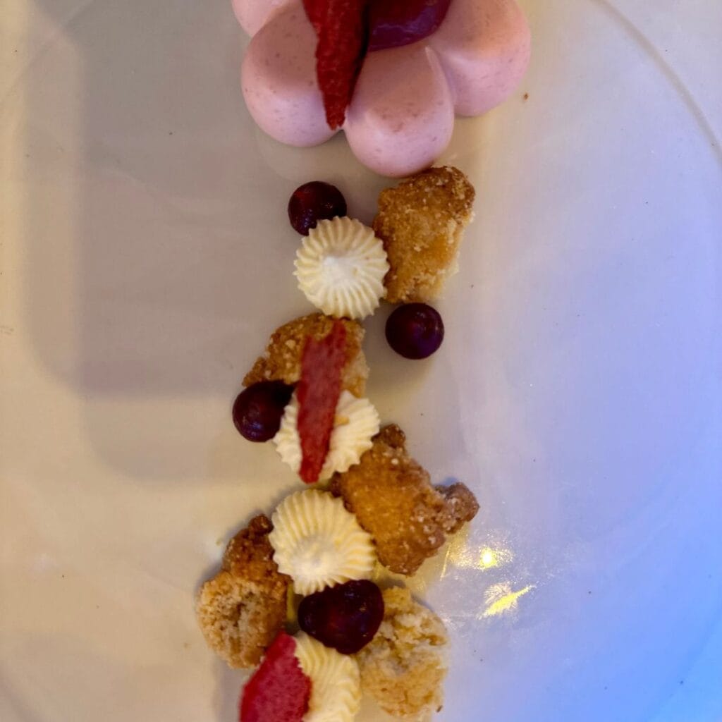 Artfully plated dessert with pink mousse topped with berry sorbet, accompanied by dollops of cream, red berries, and crispy crumbles arranged in a line.