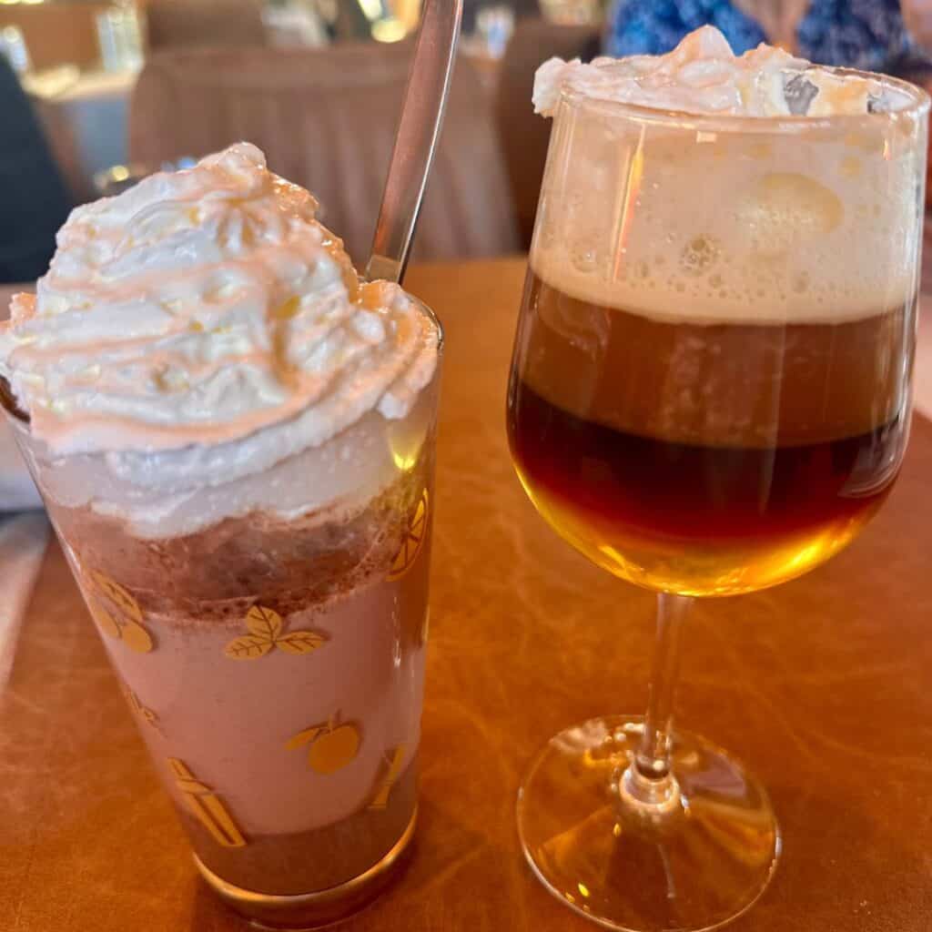 Two indulgent drinks on a restaurant table: one is a hot chocolate piled high with whipped cream, the other a layered Irish coffee with cream on top.