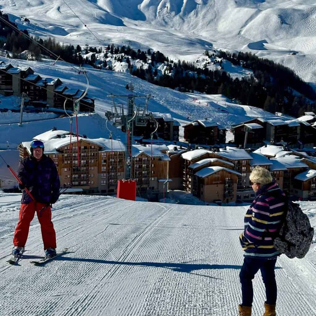 A skier in red pants poses at the top of a snowy slope in La Plagne while another person in a striped jacket stands nearby, with a ski lift and snow-covered alpine village in the background.