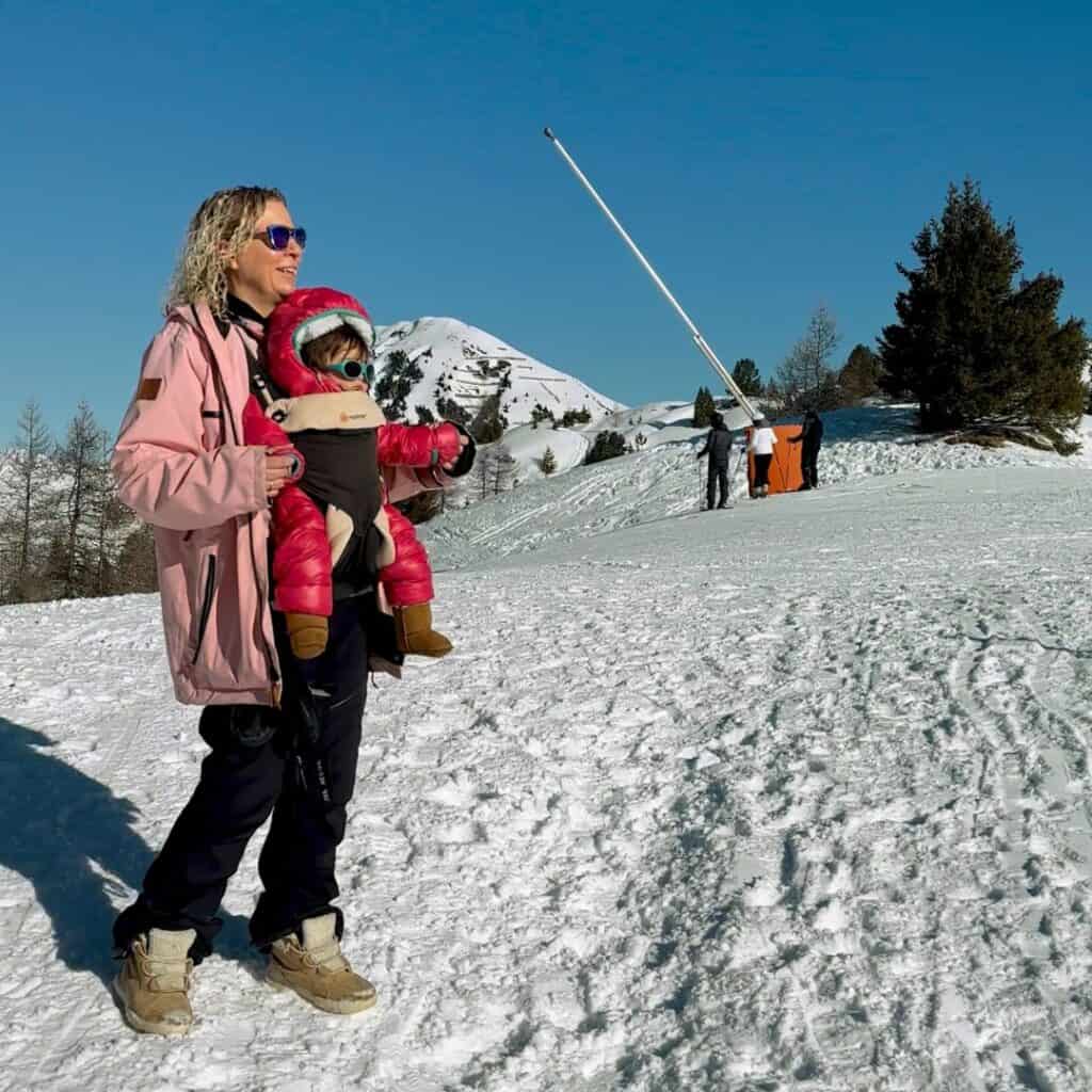 A woman in a light pink ski jacket stands on a snowy mountain path, wearing sunglasses and carrying an infant in a front-facing baby carrier. Both are warmly dressed and enjoying the sunny alpine view.